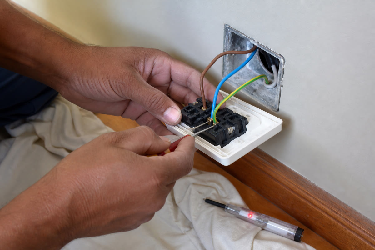 Hands wiring a South African 16A wall socket with brown live, blue neutral, and earth conductors, voltage tester on the floor.